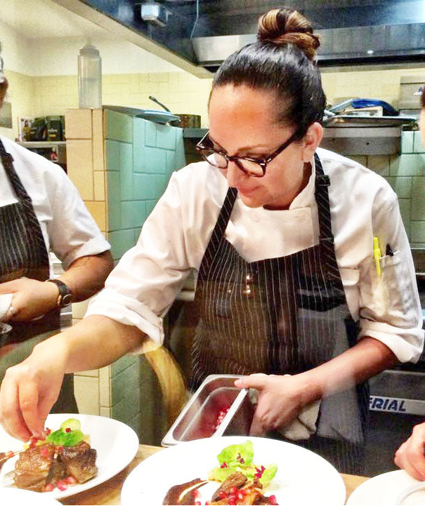 Chef Lori Sandoval in a restaurant Los Angeles kitchen preparing food, wearing an apron and glasses.