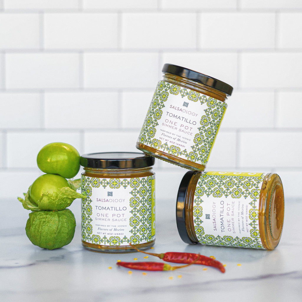 Three jars of tomatillo sauce with decorative labels on a white surface with tomatillos and red peppers.