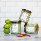 Three jars of tomatillo sauce with decorative labels on a white surface with tomatillos and red peppers.